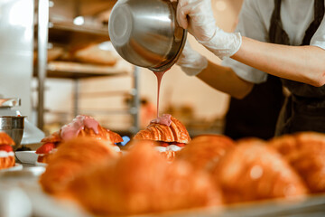 Confectioner pours pink icing onto tasty croissant from metal bowl on counter