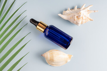 A glass bottle with face oil or serum on a blue background with seashels and tropical palm leaf....