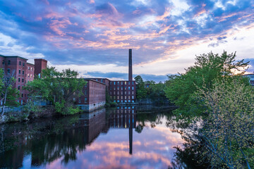 Historic cotton mill building with a tall brick chimney on the banks of the river in the old Nashua Industrial Park and its reflection in the Nashua River. New Hampshire, USA