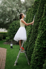 beautiful young woman gardener in a dress cuts pyramidal thujas from a stepladder, topiary art and landscaping in the garden, a classic garden and a girl in a beautiful puffy dress