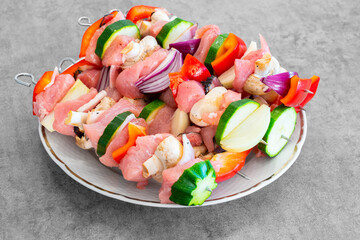 Raw turkey shish kebab on metal skewers, with lots of colorful veggies, on a white plate, ready for preparation. Light grey design desk surface. Delicious, light, great looking food.