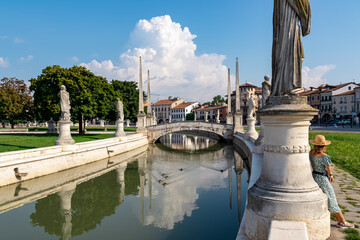 Naklejka premium Woman in a dress and hat leaning against columns of a statue at Prato della Valle, square in the city of Padua, Veneto, Italy, Europe. Isola Memmia surrounded by canal bordered by two rings of statues
