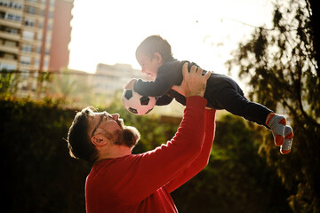 Man having a good time with his little son while holding him up in the air with a soccer ball.