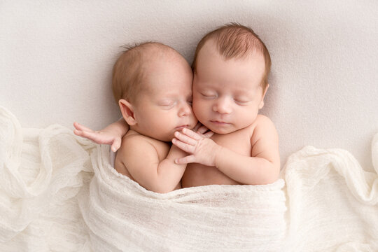 Tiny newborn twins boys in white cocoons on a white background. A newborn twin sleeps next to his brother. Newborn two twins boys hugging each other.Professional studio photography