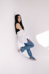 Pretty woman barefoot in studio. Beautiful brunette girl in black topic with white shirt and blue denim jeans. White background of cyclorama