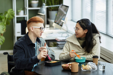Side view portrait of two smiling young people eating lunch at work and enjoying conversation