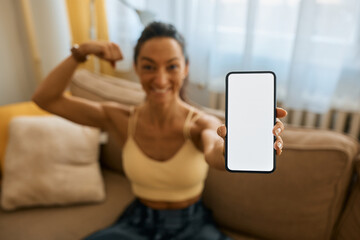 Close up of athletic woman holding smart phone with blank screen while flexing her muscles at home.