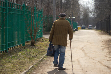 Pensioner in Russia. Poverty in world. Elderly man walks leaning on walking stick. Background man...