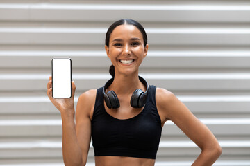 Glad young arab lady athlete with headphones shows smartphone with blank screen on wall background