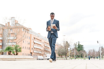 African American business man walking and holding tablet pc and cup of coffee. Young man in suit outdoors. Breaks increase productivity at work.