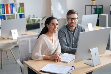 Happy young woman and her male colleague working together, using modern computer in open space...