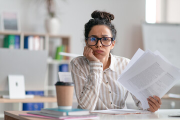 Tired female office worker looking at camera and holding documents, sitting at desk in office