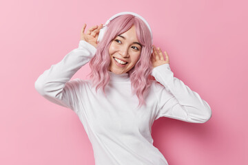 Cheerful dreamy young Asian woman looks away smiles gladfully wears wireless headphones on ears...