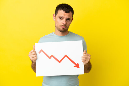 Brazilian man over isolated purple background holding a sign with a decreasing statistics arrow symbol with sad expression