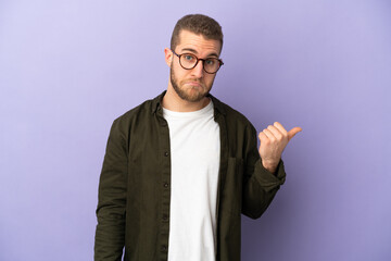 Young handsome caucasian man isolated on purple background unhappy and pointing to the side