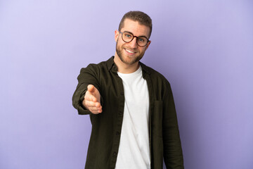 Young handsome caucasian man isolated on purple background shaking hands for closing a good deal