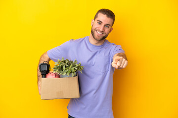 Young caucasian making a move while picking up a box full of things isolated on yellow background...