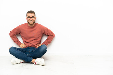 Young handsome man sitting on the floor posing with arms at hip and smiling