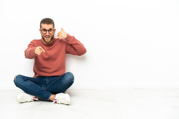 Young handsome man sitting on the floor making phone gesture and pointing front
