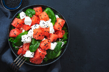 Summer Watermelon juicy salad bowl with feta cheese, spinach and black sesame seed, dark table background, top view, negative space