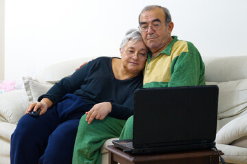 Happy elderly couple are using laptop while talking sitting on sofa at home. they shop on the internet, plan a family vacation, surf the internet, laugh, have fun together with the computer.