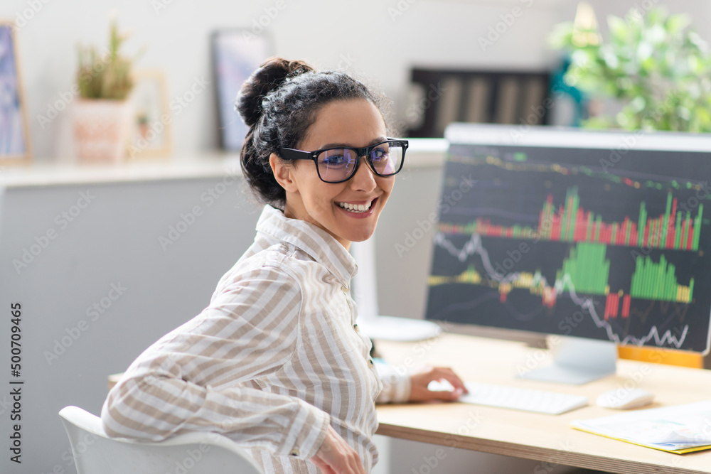 Poster happy businesswoman sitting at workplace in office and smiling at camera, computer monitor with grap