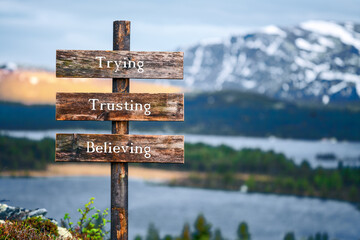 trying trusting believing text quote written on wooden signpost outdoors in nature with lake and mountain scenery in the background. Moody feeling.