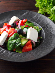 Fresh Greek salad with tomatoes, cucumbers, bell peppers, olives, purple onions and feta cheese on a black plate, top view, dark wooden background. Vegan salad with tofu cheese. Copy space. Close up.