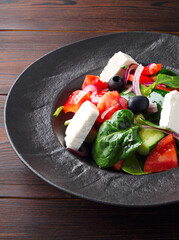 Fresh Greek salad with tomatoes, cucumbers, bell peppers, olives, purple onions and feta cheese on a black plate, top view, dark wooden background. Vegan salad with tofu cheese. Copy space. Close up.