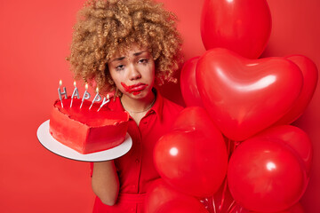 Curly haired woman has spoiled makeup looks frustrated has unhappy expression feels lonely during holiday holds sweet cake and heart shaped balloons isolated over red background. Festivity concept
