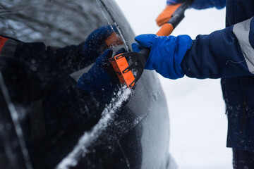 A human hand in a glove cleans a blue car from snow with a special brush. A hand sweeps snow from the hood of the car. Horizontal photo. the consequences of a blizzard