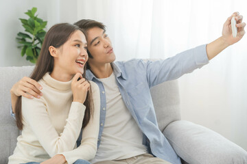 Fototapeta na wymiar Happy couple love at home, two asian young spending time, bonding to each other romantic on sofa in living room while man embrace woman using smartphone, mobile phone to selfie take a photo together.