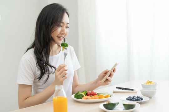 Dieting, asian young woman eating, holding fork at broccoli, diet plan nutrition with fresh vegetables salad, enjoy meal while using smartphone. Nutritionist of healthy, nutrition of weight loss.
