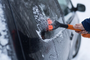 A human hand in a glove cleans a blue car from snow with a special brush. A hand sweeps snow from...