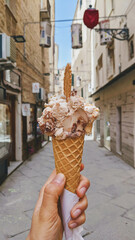 Mano che regge un cono gelato vegano alle creme con cialda biscotto in una via di Alghero, Italia...