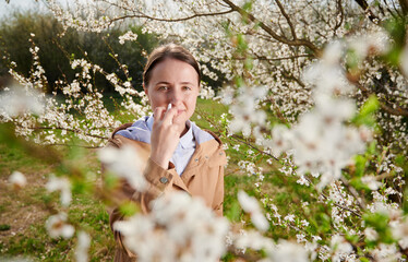 Woman allergic suffering from seasonal allergy at spring, posing in blossoming garden at...