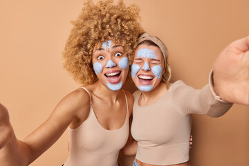 Cheerful women pose for making selfie keep arms outstretched apply beauty mask on face have fun embrace and stand closely to each other giggle happily isolated over brown background. Skin care