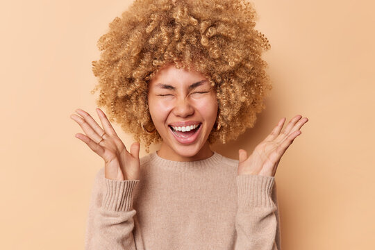 Cheerful optimistic woman keeps palms raised up yells happily closes eyes smils broadly shows white teeth dressed inn casual jumper isolated over brown studio background. Human reactions concept
