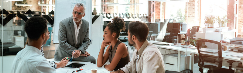 Working process. Mature businessman talking about something with his young colleagues while sitting together at the office table in the modern office