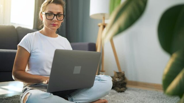 Woman with glasses is sitting on the floor and working on a laptop. Cat lies in the background. Concept of remote work.