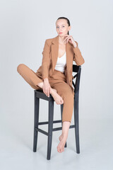 Serious woman in a beige suit. Sits on a high wooden black chair with bare feet. Full length portrait. Isolated on white background.