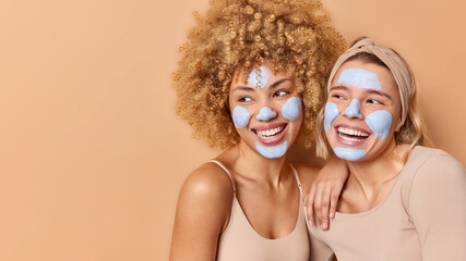 Horizontal shot of positive young women apply nourishing beauty masks for face care look gladfully away undergo skin treatments isolated over brown background copy space for your advertising content