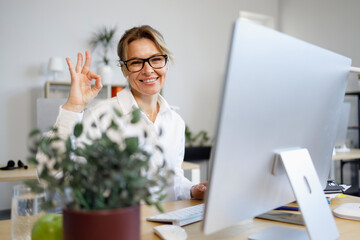 Middle-aged business woman sits at a computer in the office and shows ok gesture with her fingers