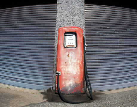 Alte, rote, rostige Benzin-Zapfsäule vor neutralem Garagen-Rolltor. Ausser Betrieb, Öl-Embargo.