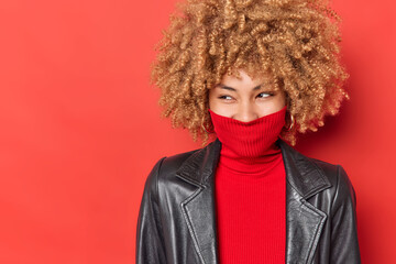 Cheerful curly haired woman looks away happily wears turtleneck covers mouth with collar dressed in black leather jacket isolated over vivid red background empty space for your advertisement