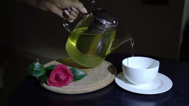 Glass teapot and red camellia on a wooden stand with green tea in the dark. A beautiful female hand appears and pours tea into a cup. Fancy black background