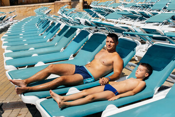 A young athletic man and his son are smiling happily and sunbathing on a sun lounger on a sunny day...