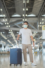 Young male wearing face mask with luggage walking in airport, protection Coronavirus disease infection, Asian man traveler with hat. Time to travel after vaccine booster dose concept
