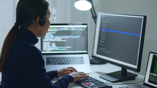 Asian woman developer programmer, software engineer, IT technical support wearing headsets talking with team, working overtime at night on computer to check coding bugging system. Back view