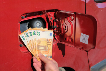 A hand holding a bundle of fifty euro notes in font of a fuel tank.